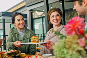 A group enjoying dinner on a patio, one woman is passing a plate with a fish burger to a man.